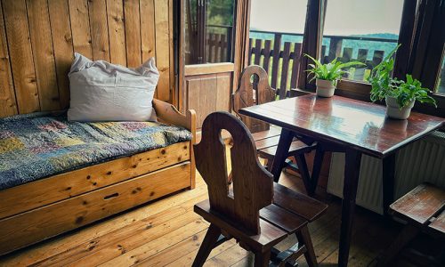 drewniany pokój w schronisku, widać łózko z poduszką, otwarte drzwi na balkon, drewniany stolik i kwiaty w doniczkach