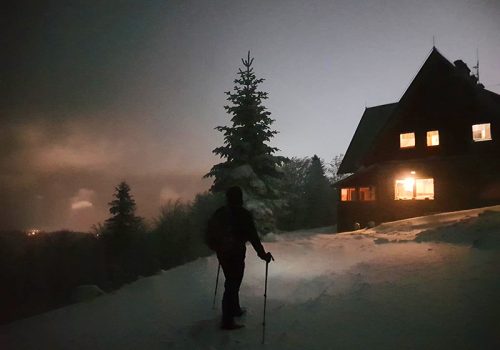 schronisko górskie nocą. wokół leży śnieg, świtało pada na turystę, który stoi na nartach
